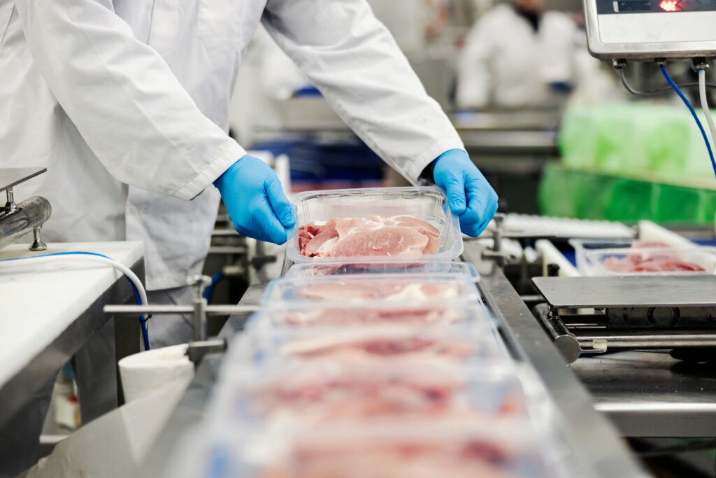 Person in white coat and latex gloves examining packaged meat on a conveyor belt.