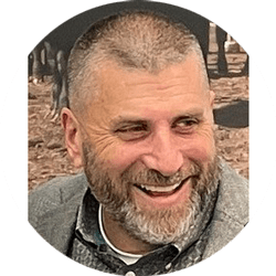 Smiling man with short gray beard and cropped hair, wearing a gray jacket, outdoors in a park.