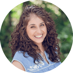 Portrait of a smiling woman with curly brown hair outdoors, wearing a light blue embroidered top.