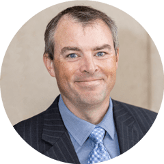 Head-and-shoulders portrait of a smiling man in a dark pinstripe suit, light blue shirt, and patterned tie.