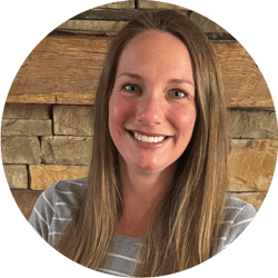 Smiling woman with long light brown hair, wearing a gray striped shirt, in front of a wooden log background (circular crop).