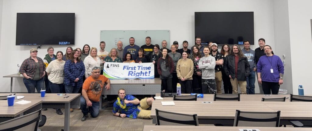 Group of people in a classroom posing for a photo behind a banner reading'First Time Right!'