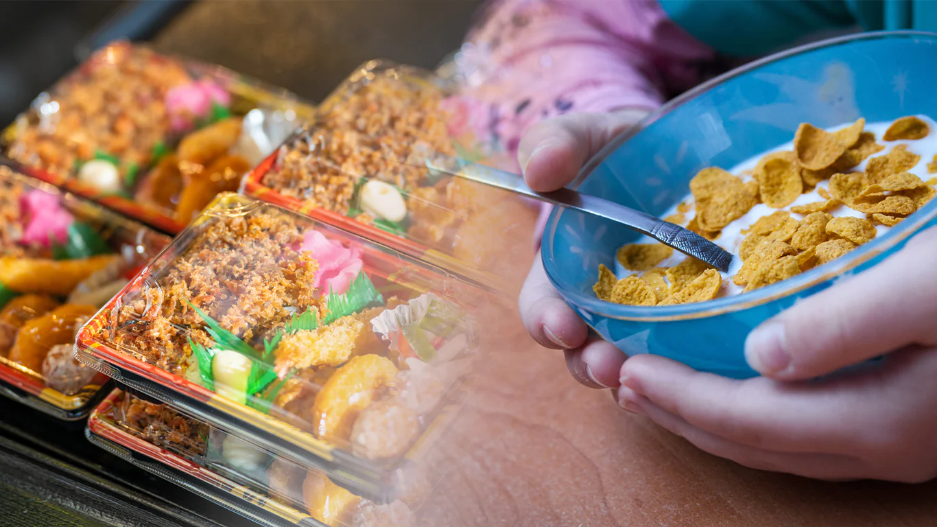 Person holding a blue bowl of cornflakes with milk and a spoon, near a display of colorful bento boxes on a wooden table