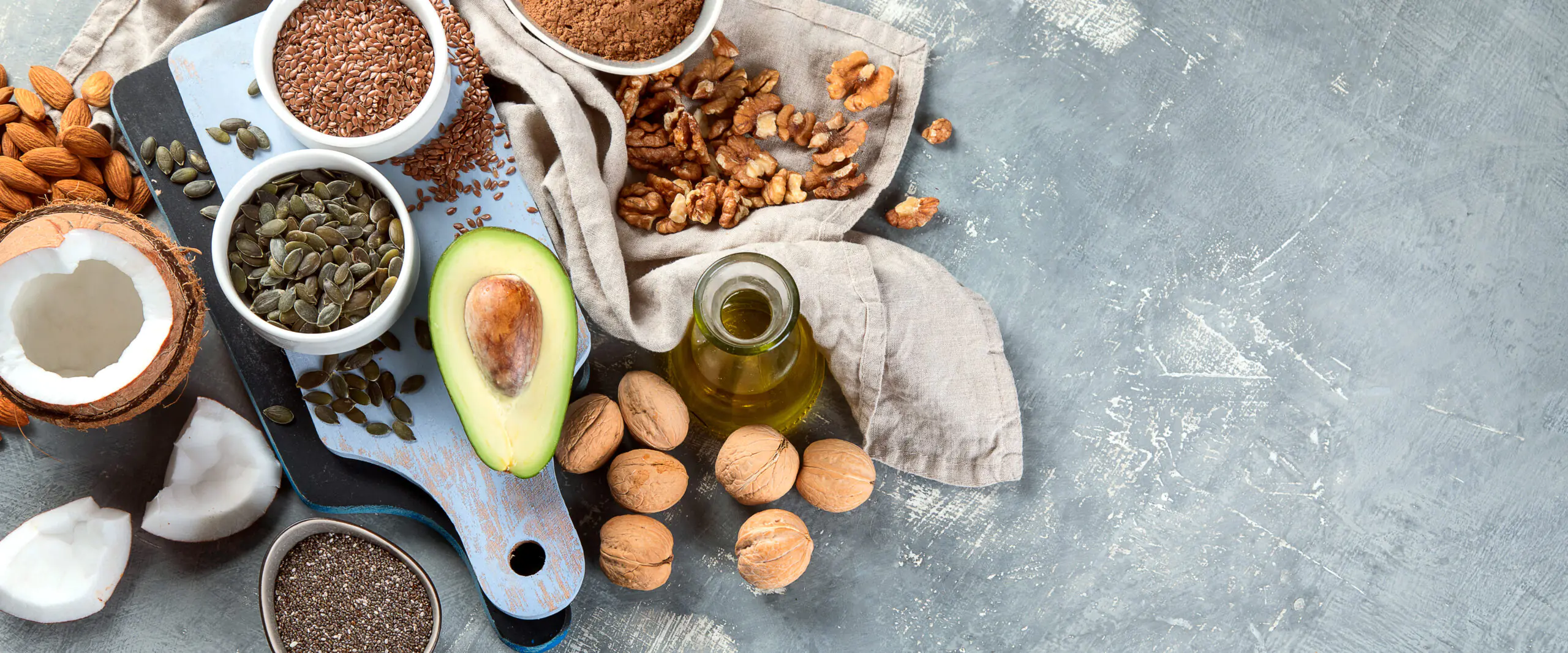 Flat lay of almonds, walnuts, seeds, coconut, avocado half, and olive oil on a blue-gray surface.